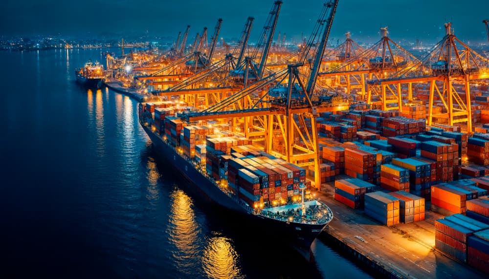 A nighttime aerial view of a busy port filled with illuminated cranes and cargo ships, surrounded by stacked shipping containers. 