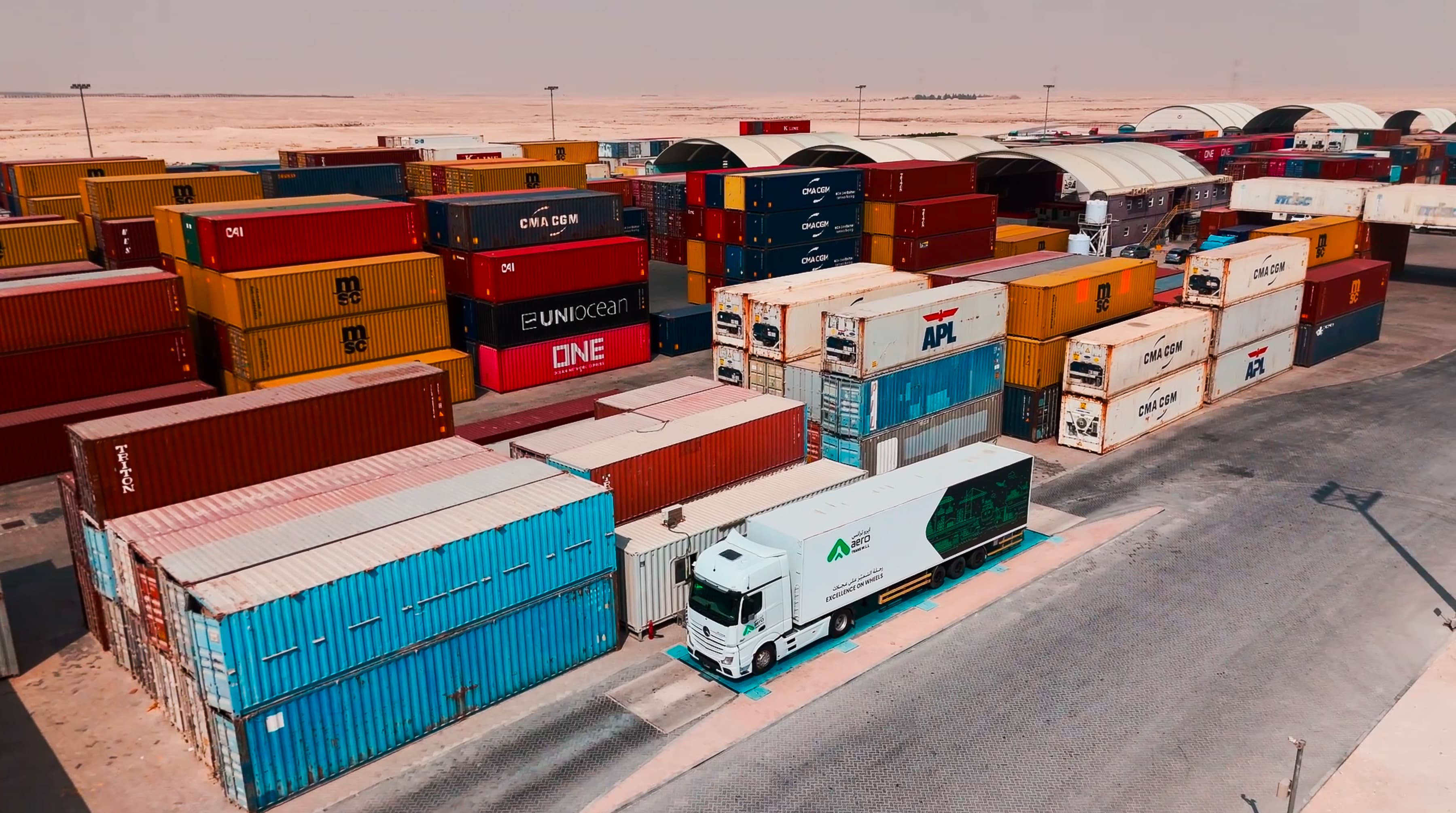 A truck navigates between stacked colorful shipping containers under a clear sky in a desert-like industrial area, conveying a sense of logistics and commerce. 