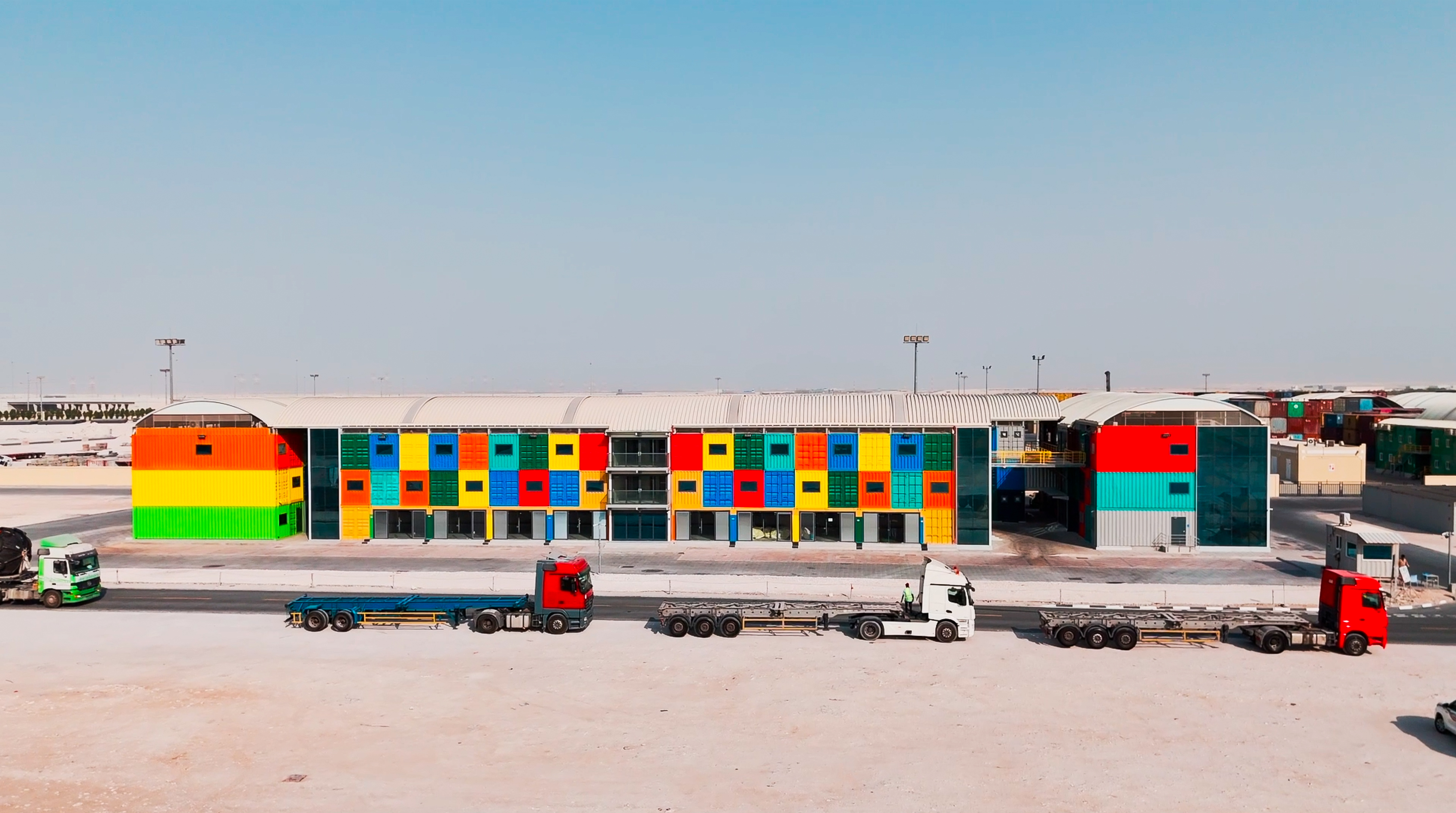 A vibrant, modern building with a colorful facade of red, blue, green, and yellow panels. Four large trucks are parked in front under a clear sky