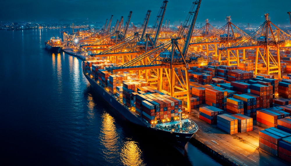 A nighttime aerial view of a busy port filled with illuminated cranes and cargo ships, surrounded by stacked shipping containers.