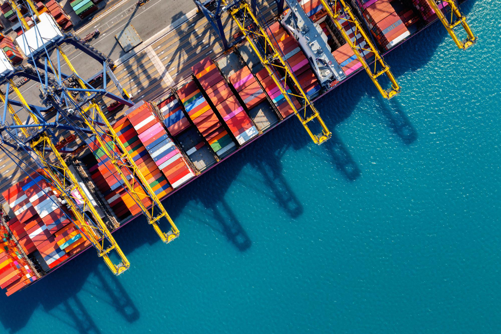 Aerial view of a port with colorful shipping containers and cranes casting shadows on turquoise water.